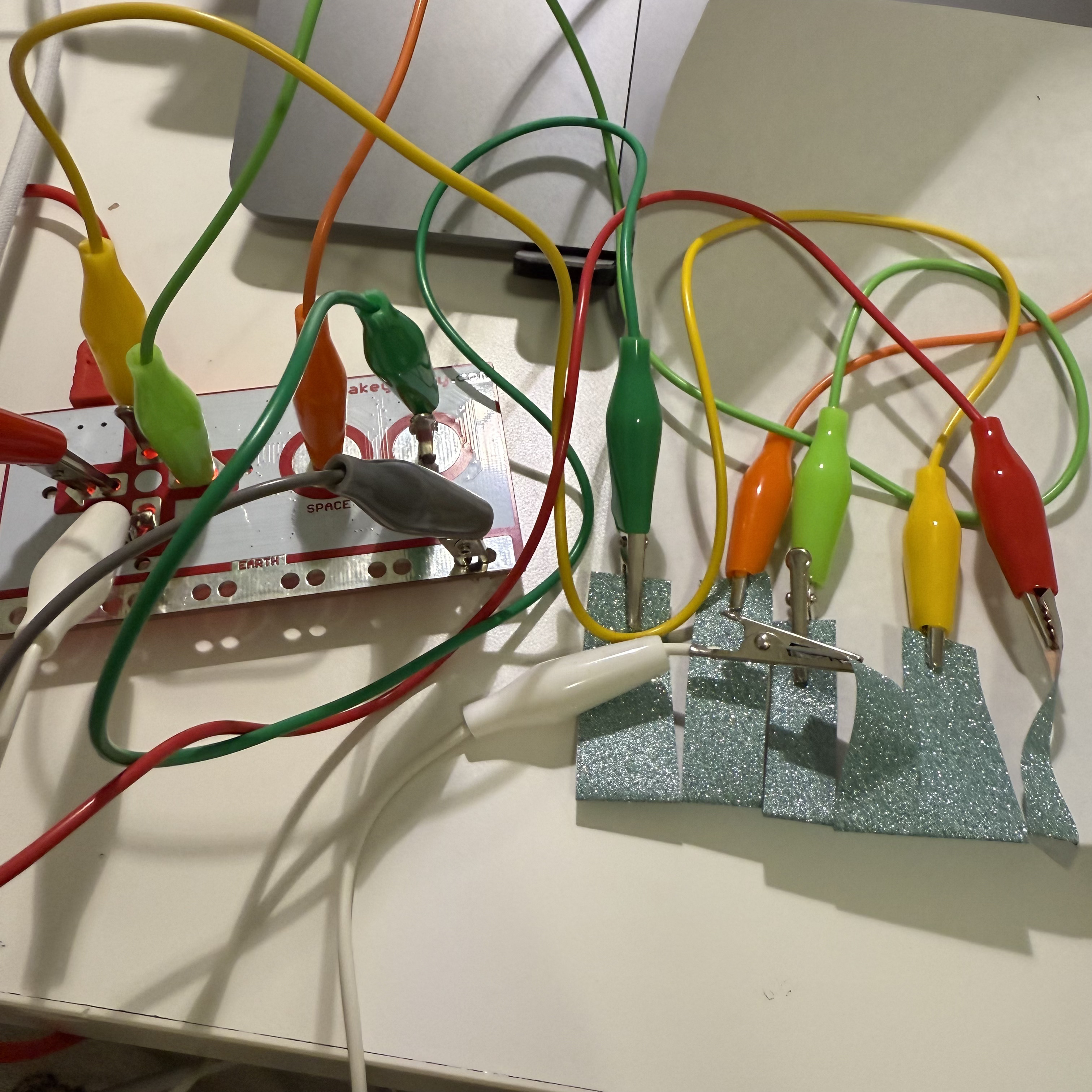 On a desk, the Makey Makey setup is connected to a laptop, which displays a colorful interactive game. When the conductive materials are touched, they control the game shown on the laptop screen, demonstrating how physical interaction can manipulate digital environments.